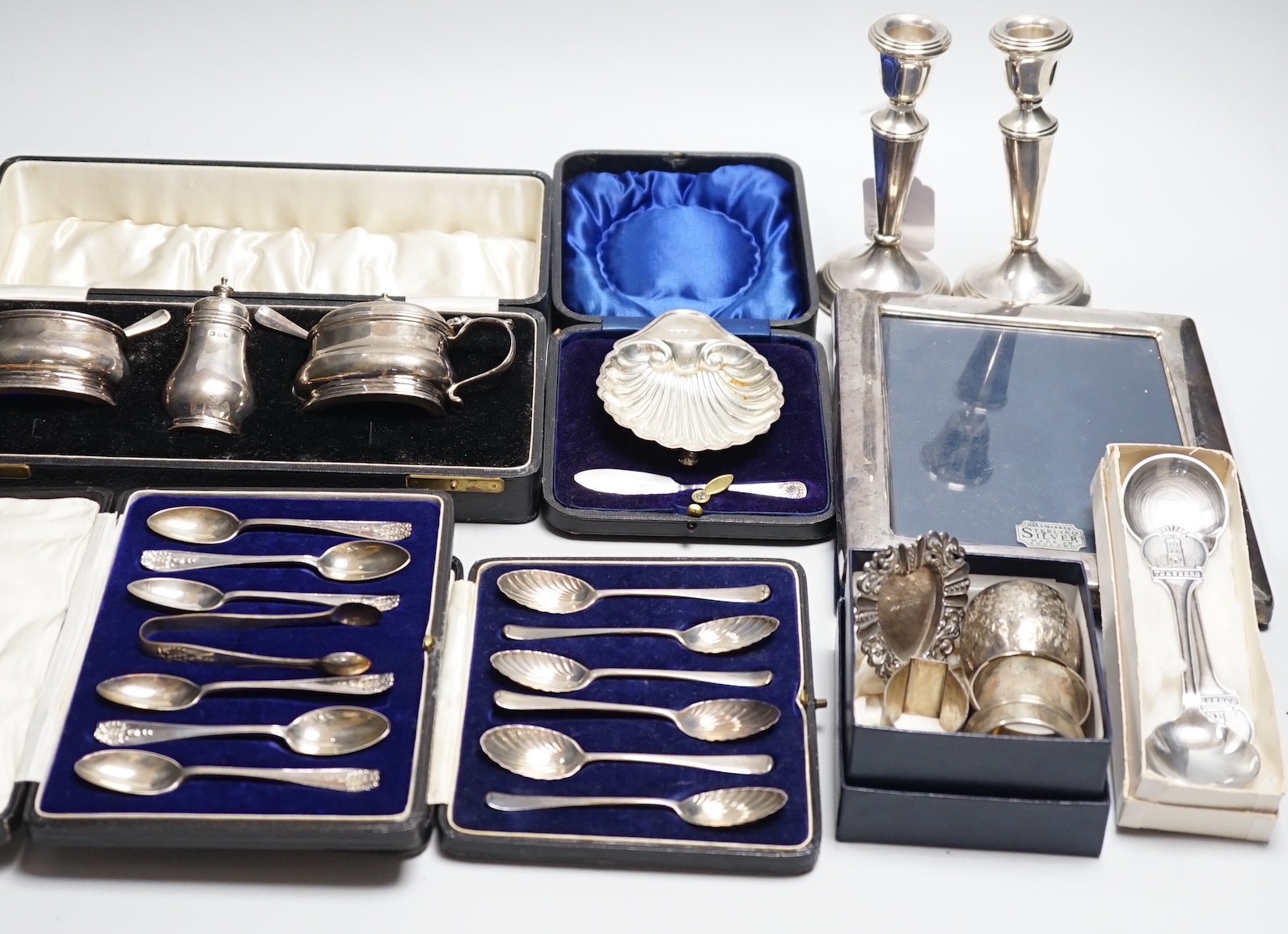 A quantity of mixed silver ware including a pair of modern candlesticks, 15cm, weighted, a cased George V three piece condiment set, cased butter shell and knife, two cased sets of teaspoons, a mounted photograph frame,